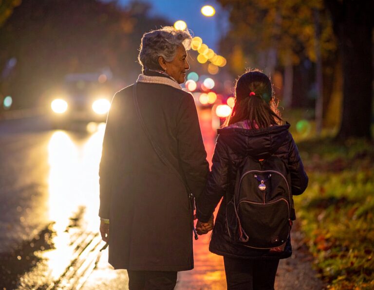 Starsza osoba i dziecko idące wieczorem za rękę wzdłuż mokrej ulicy, oświetlonej reflektorami samochodów i miejskimi latarniami.