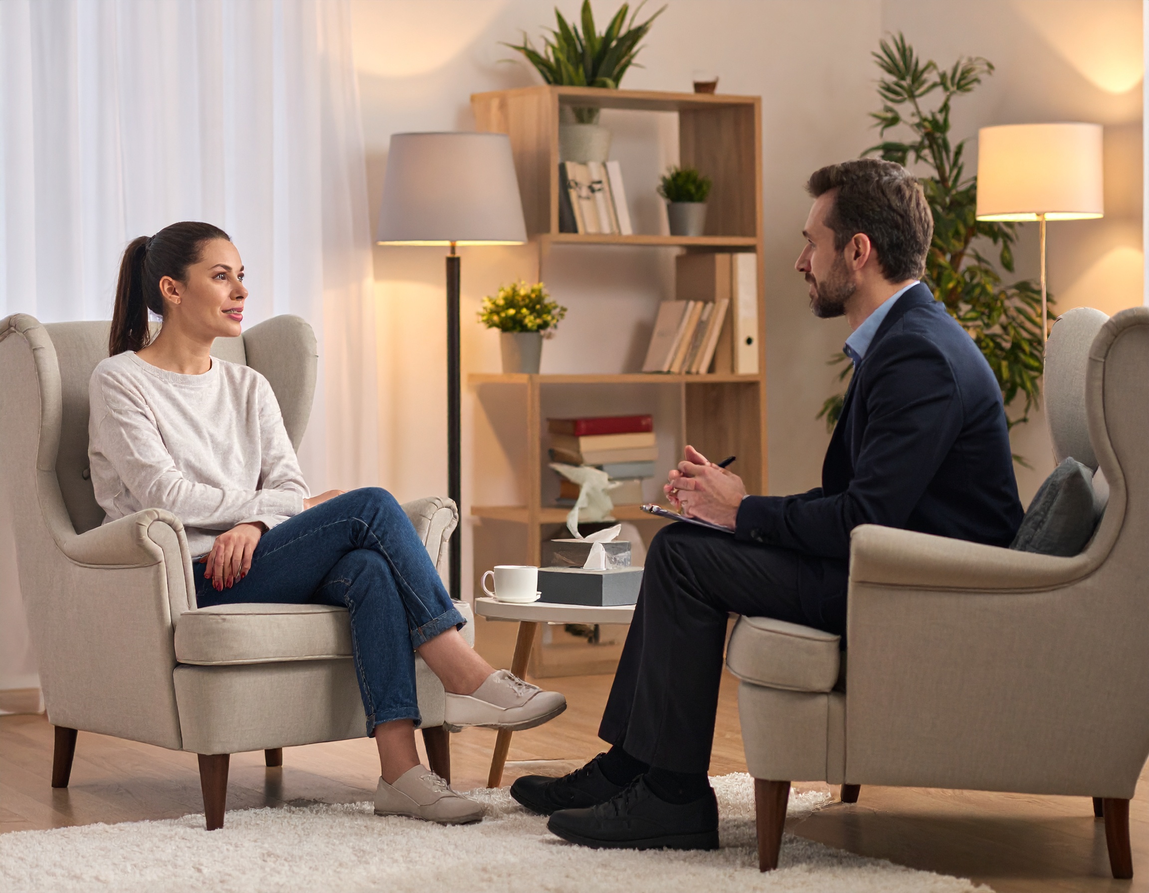 Kobieta rozmawiająca z terapeutą w przytulnym gabinecie, siedzą na fotelach naprzeciwko siebie, w tle regał z książkami i roślinami.