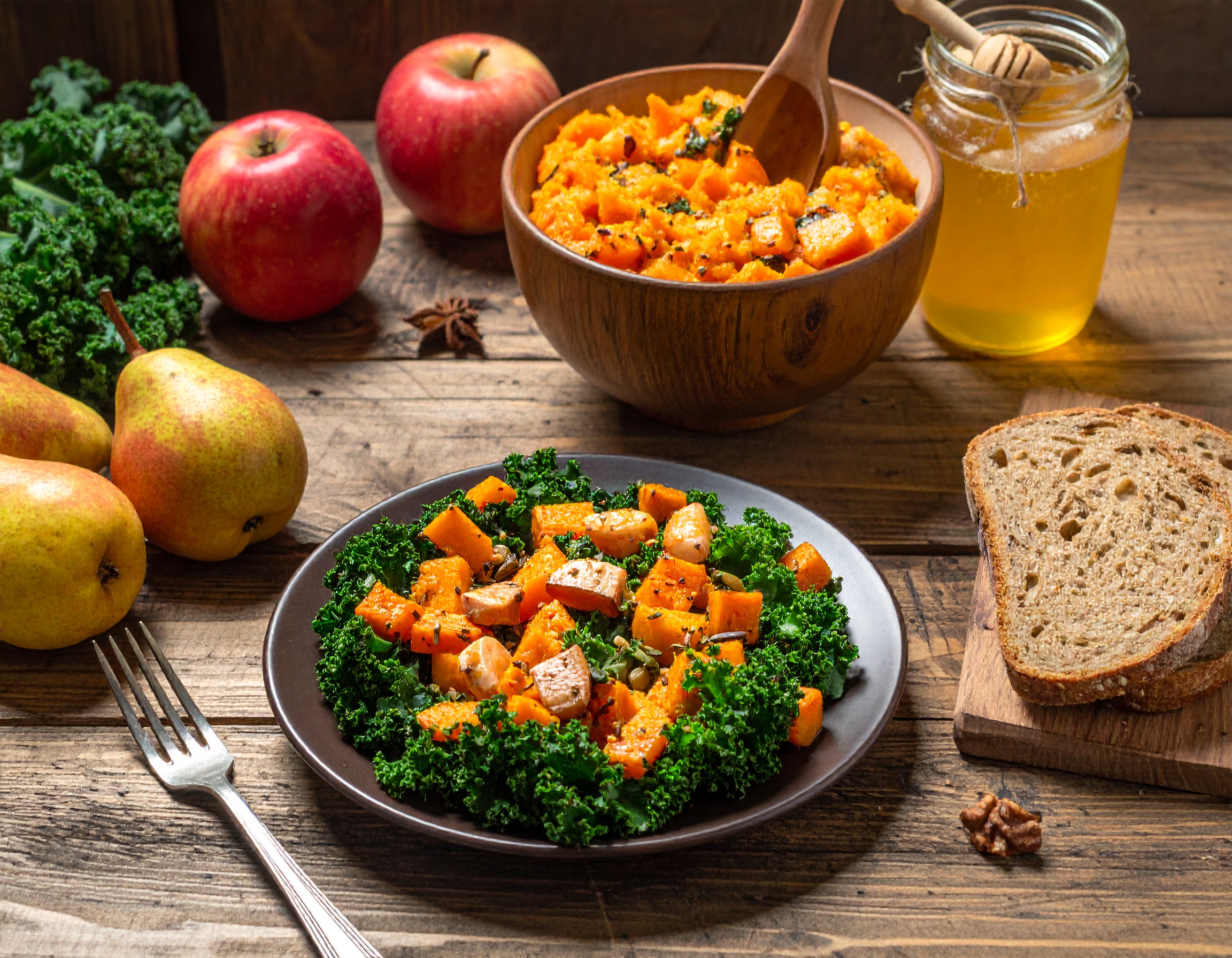 Jesienne danie z pieczonej dyni i jarmużu podane na talerzu, obok gruszki, jabłka, chleb pełnoziarnisty i słoik miodu na drewnianym stole.