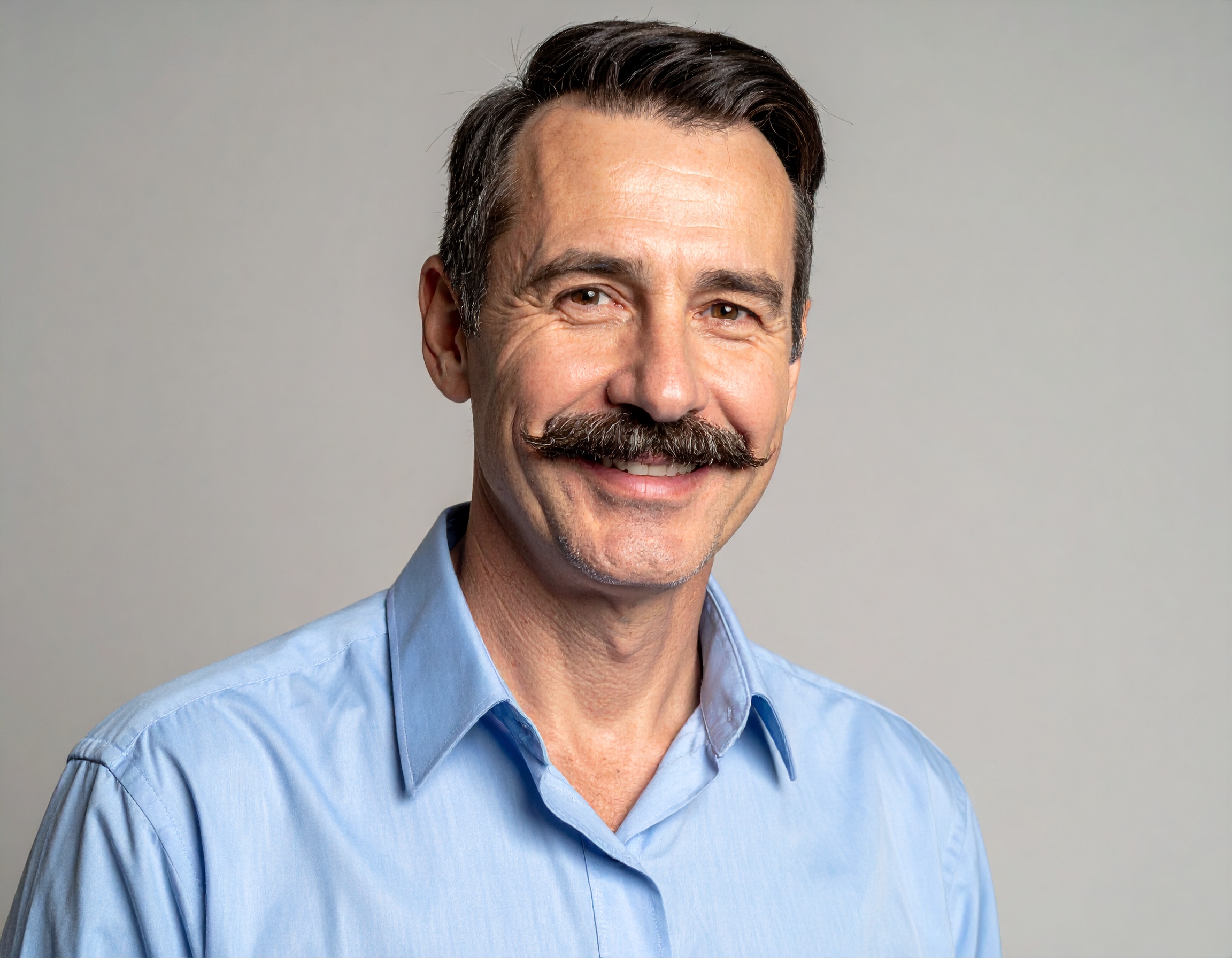Uśmiechnięty mężczyzna w średnim wieku z wąsem, ubrany w niebieską koszulę, na neutralnym tle.