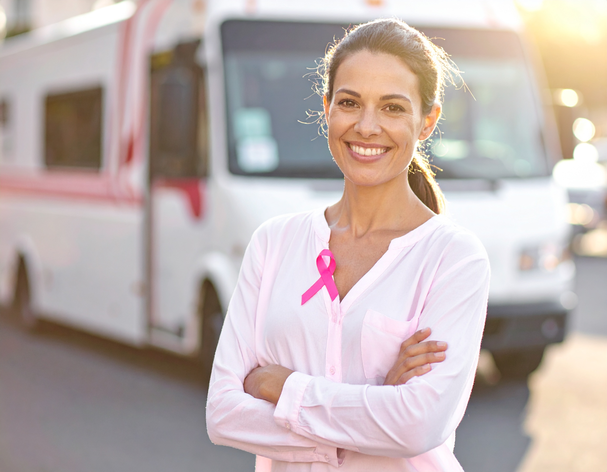 Uśmiechnięta kobieta w jasnoróżowej bluzce z przypiętą różową wstążką, stojąca przed mobilnym punktem medycznym.