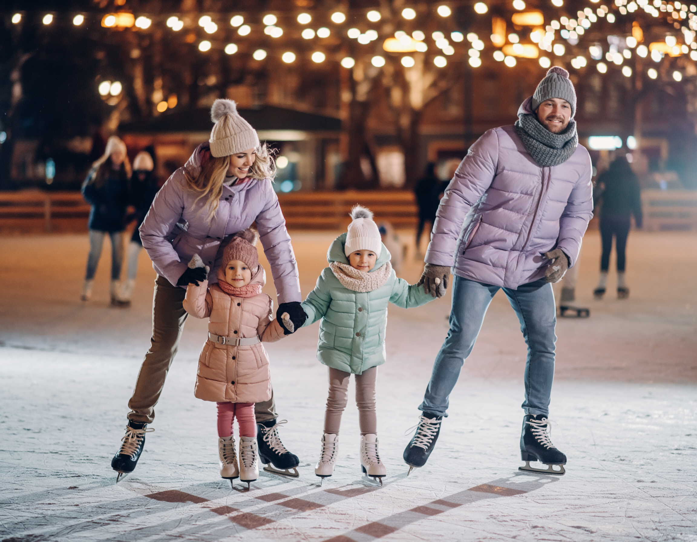 Rodzina z dwójką dzieci jeżdżąca wspólnie na łyżwach na oświetlonym, zimowym lodowisku wieczorową porą.