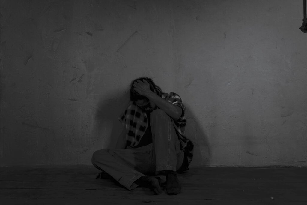 Osoba siedząca na podłodze w ciemnym pomieszczeniu, z głową opartą na dłoni, wyglądająca na przygnębioną i przytłoczoną.