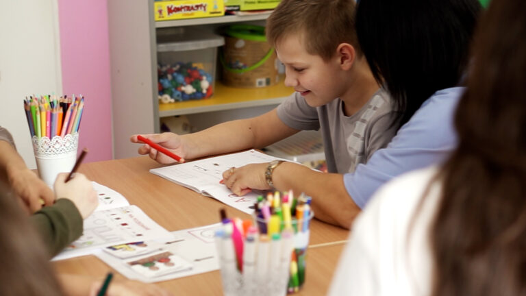 Chłopiec pracuje przy stoliku z terapeutką, wykonując ćwiczenia w zeszycie, obok stoją kolorowe kredki i pomoce edukacyjne.