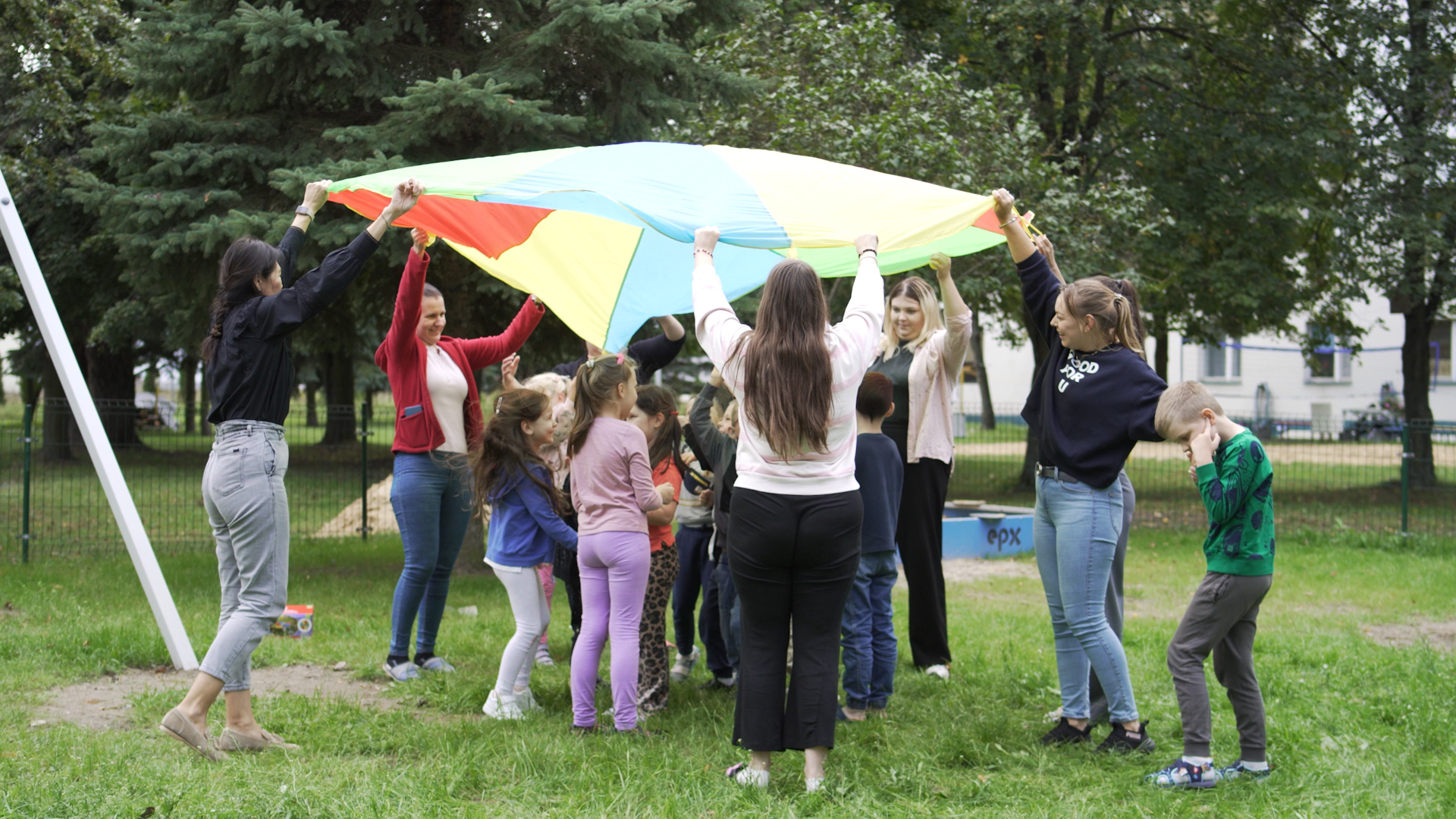 Grupa dzieci i dorosłych bawi się wspólnie na trawie, trzymając kolorową chustę animacyjną podczas zajęć integracyjnych.