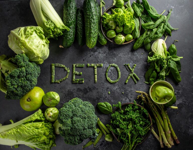 Napis „DETOX” ułożony z zielonych warzyw na ciemnym tle, otoczony brokułami, ogórkami, sałatą, szparagami i szpinakiem.
