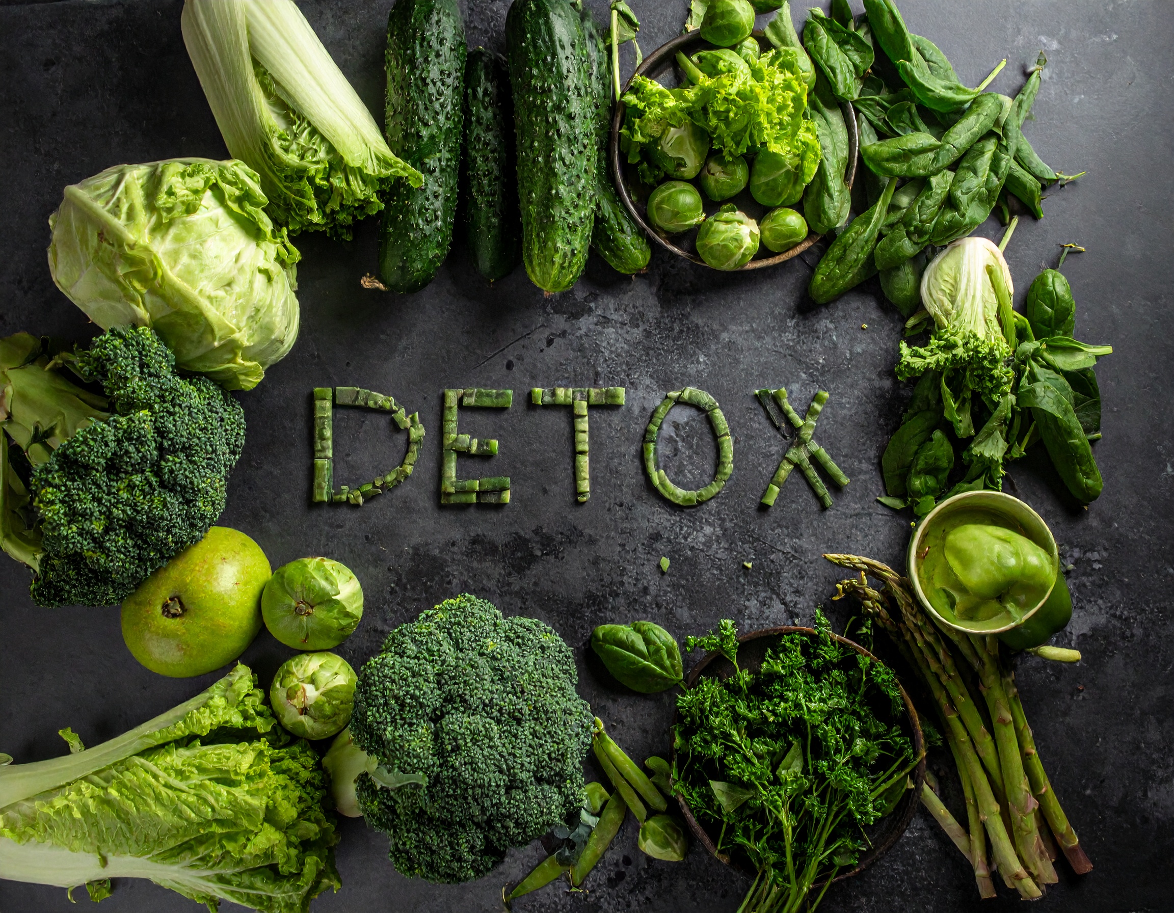Napis „DETOX” ułożony z zielonych warzyw na ciemnym tle, otoczony brokułami, ogórkami, sałatą, szparagami i szpinakiem.