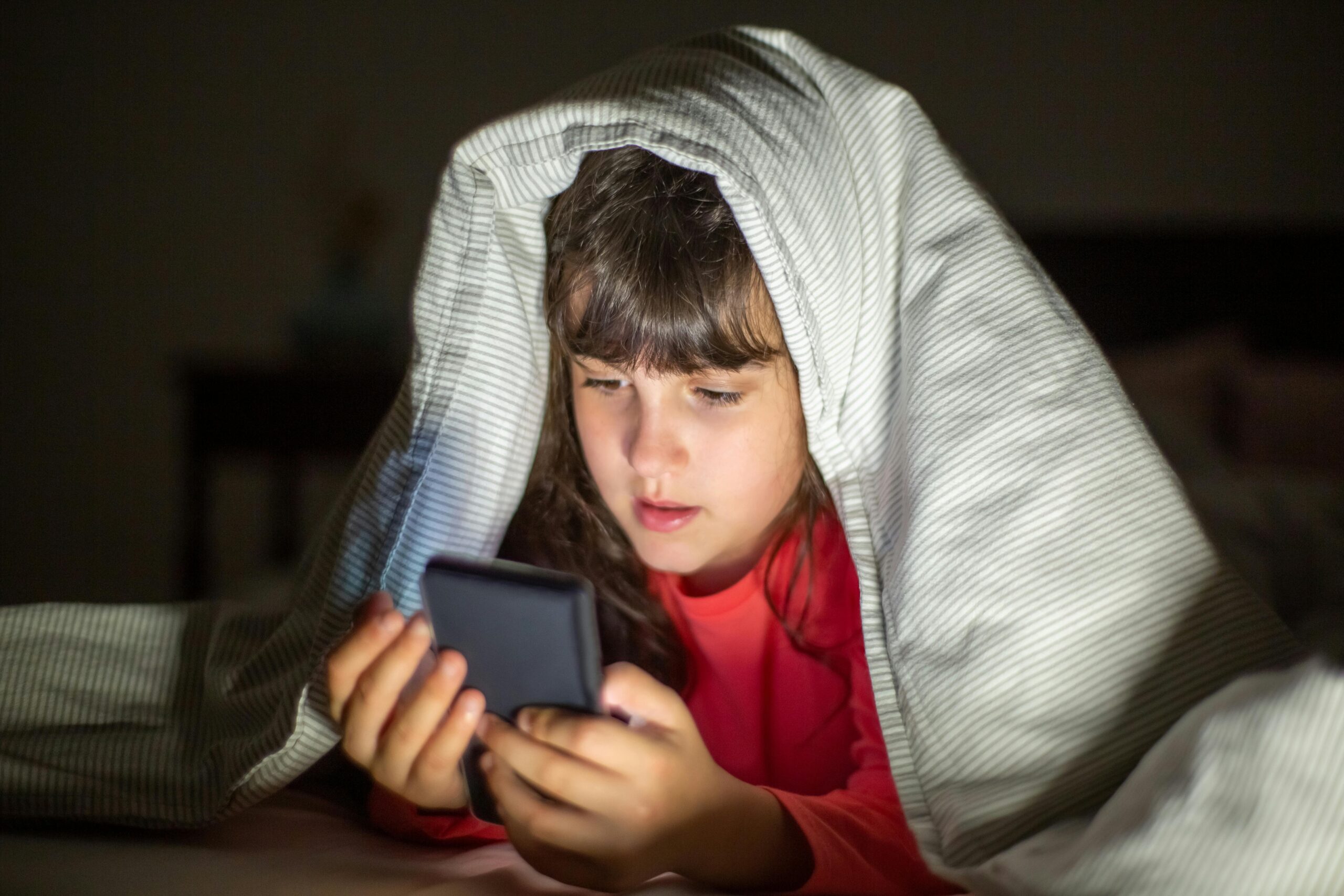 Dziecko korzysta ze smartfona późnym wieczorem, siedząc pod kołdrą – ekran telefonu oświetla twarz w ciemnym pomieszczeniu.