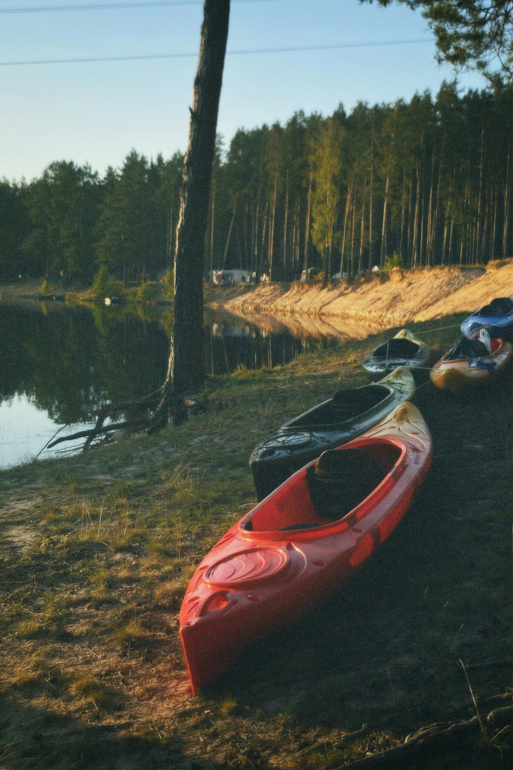Kolorowe kajaki stoją na brzegu spokojnego jeziora otoczonego lasem, przygotowane do spływu lub rekreacji na wodzie.