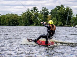 Osoba w kasku i kamizelce ochronnej pływa na wakeboardzie po jeziorze, ciągnięta przez linę na tle zielonych drzew.