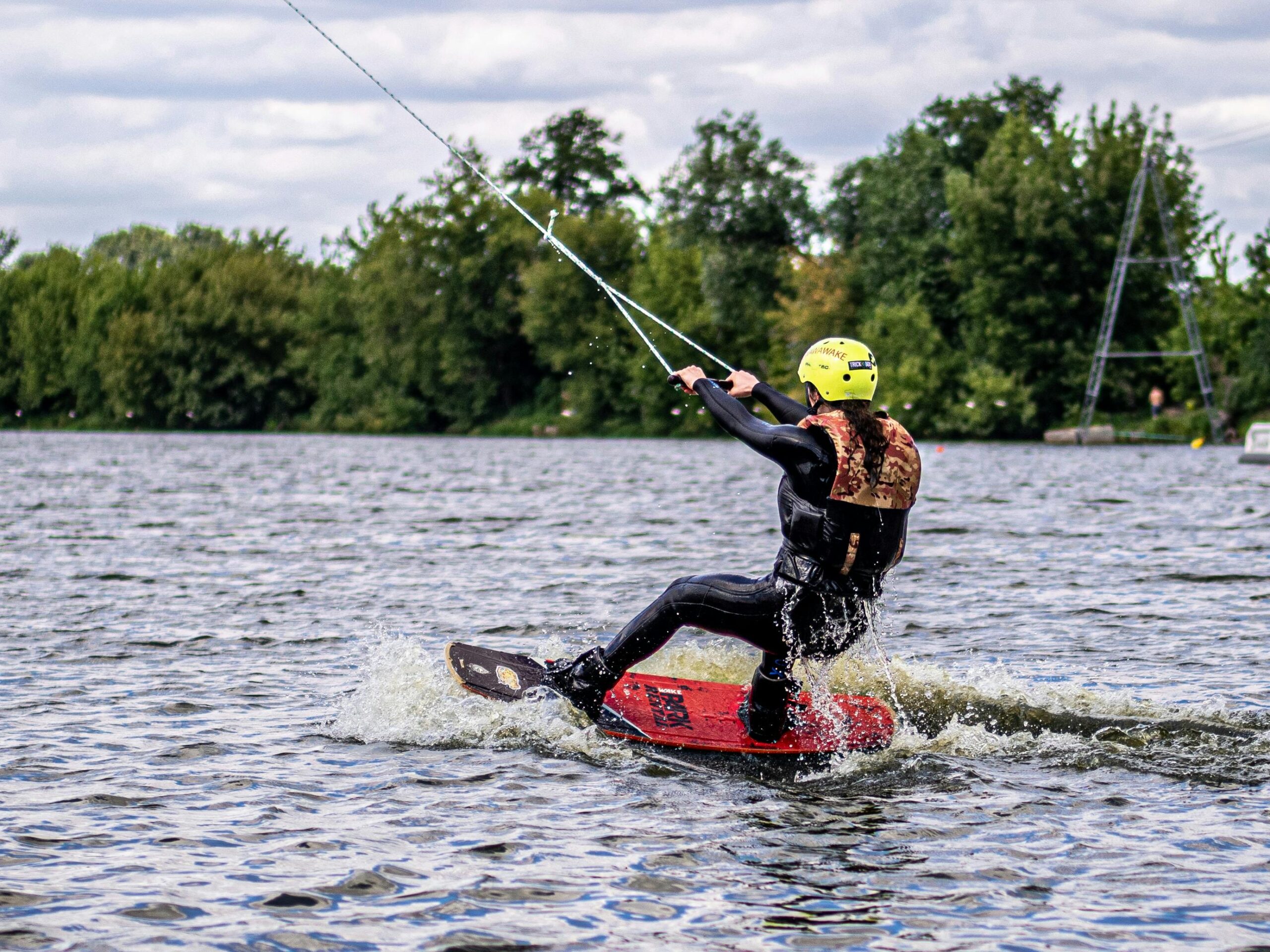 Osoba w kasku i kamizelce ochronnej pływa na wakeboardzie po jeziorze, ciągnięta przez linę na tle zielonych drzew.