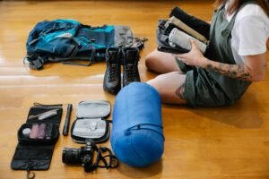 Osoba pakuje plecak i niezbędny sprzęt turystyczny przed wyjazdem w góry – obok leżą buty trekkingowe, śpiwór i akcesoria podróżne.