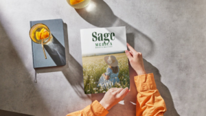 Osoba trzyma w dłoniach letnie wydanie magazynu Sage Medica na stole obok napoju i notesu.
