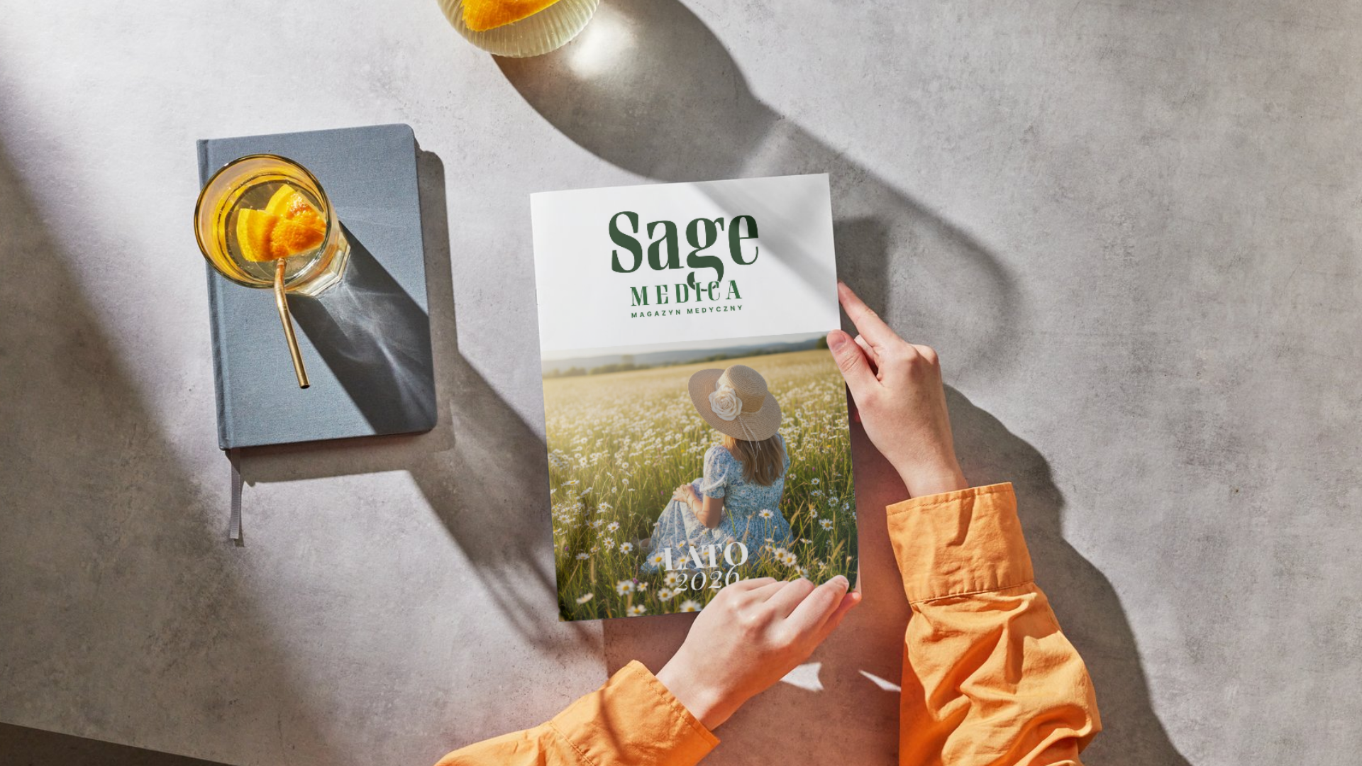 Osoba trzyma w dłoniach letnie wydanie magazynu Sage Medica na stole obok napoju i notesu.